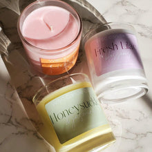 Load image into Gallery viewer, Three soy wax candles with cotton wicks, in clear glass jars, with labels indicating different fragrances such as 'Honeysuckle' and 'Fresh Lilac'. The candles are on a marble surface and one has a wooden wick.