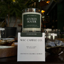 Load image into Gallery viewer, WSC Candle Co. candle with 'Stolen Hoodie' label on a stand, surrounded by decorative items.
